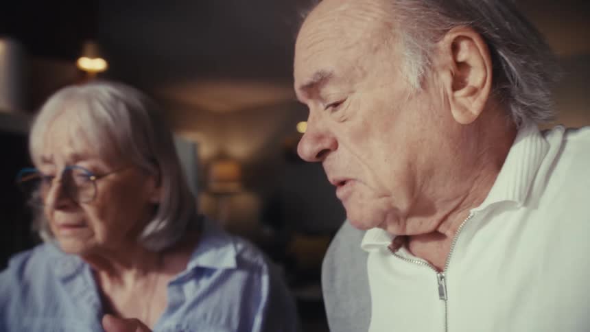 A worried elderly couple reviews their finances together, with the husband showing shock and distress after using a calculator to tally their bills