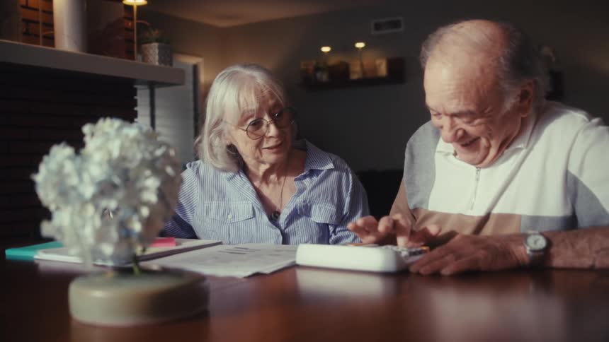 An elderly couple works on their finances together at their dining table, their initial concern turning into joyful relief as they happily review the numbers