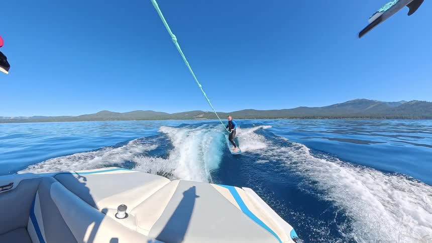 View from the back of a motorboat as a man wakesurfs across a calm blue lake, with a young girl watching from the boat on a sunny day