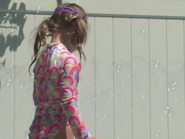 A young girl in a colorful swimsuit and goggles watches bubbles float around her in slow motion next to a kiddie pool