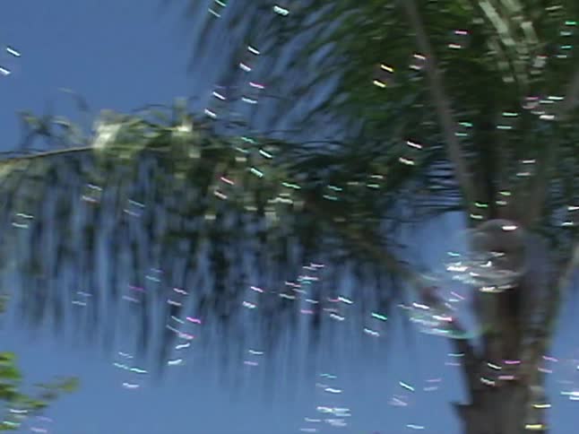A delighted little girl in goggles plays in a backyard sprinkler as the camera pans up to show bubbles floating in the sunny sky