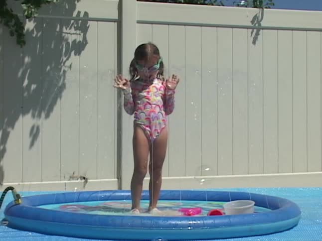 A young girl in a colorful swimsuit and goggles plays in a backyard splash pad in slow motion as bubbles float around her