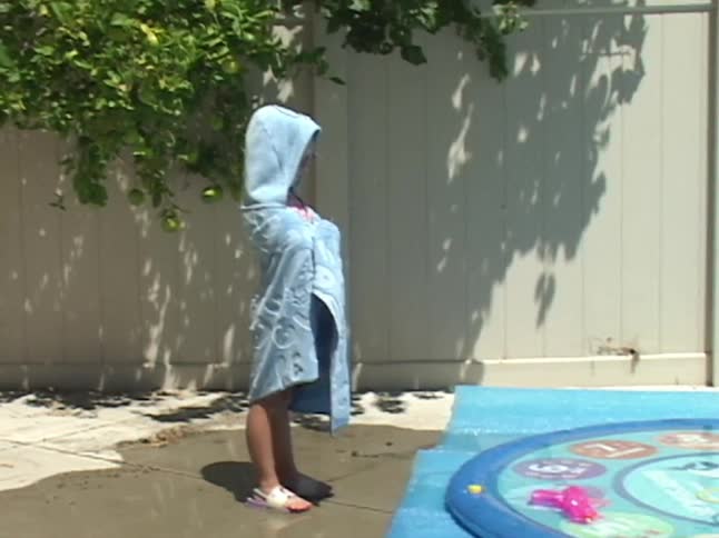 A little girl wrapped in a blue hooded towel shivers from the cold after playing in a backyard splash pad on a sunny summer day