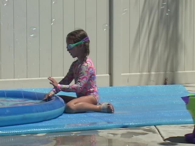 A little girl in a swimsuit and goggles plays in slow motion with bubbles from a machine, splashing in her kiddie pool on a sunny day