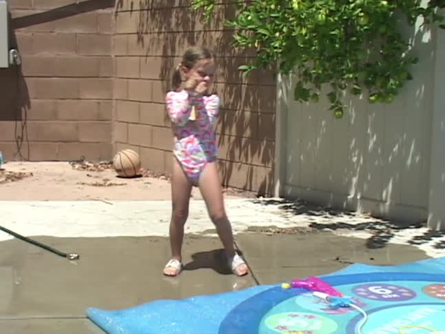 A cute little girl in a colorful swimsuit dances happily in slow motion in her sunny backyard next to a splash pad