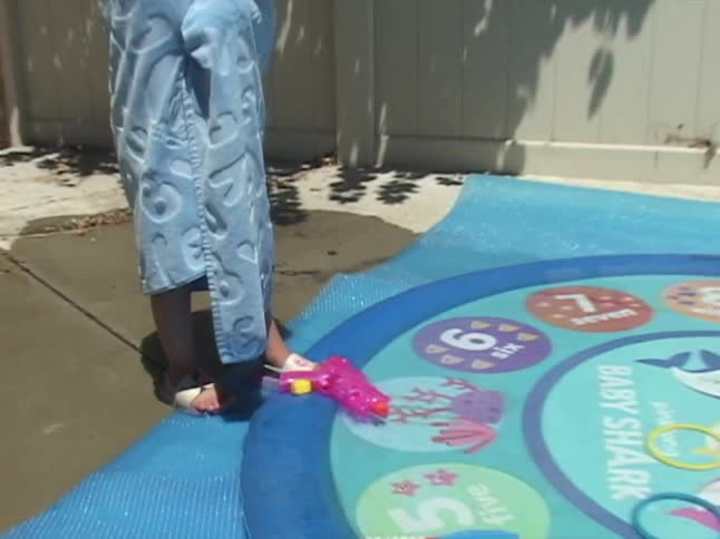 A little girl in a blue hooded towel stands shivering by her Baby Shark splash pad in the backyard on a sunny day