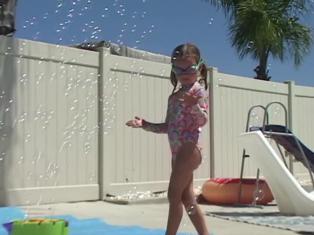 A joyful young girl in a swimsuit and goggles delightedly plays and dances as a bubble machine fills her sunny backyard with countless bubbles