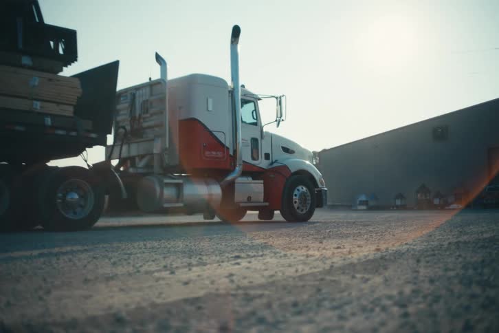 A large semi-truck loaded with lumber drives slowly through a gravel industrial lot with a cinematic sun flare