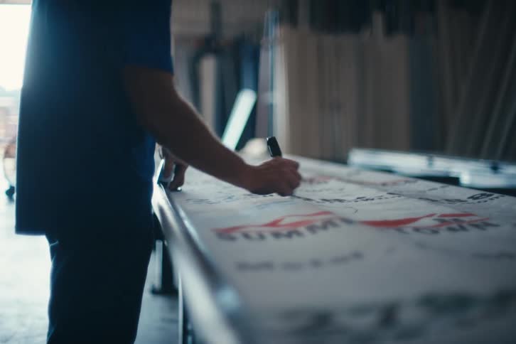 A person in a blue shirt signs their name on a large banner with a black marker in a workshop