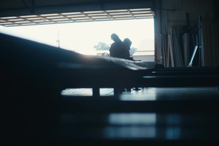 A low-angle tracking shot reveals three silhouetted workers collaborating in a large industrial warehouse