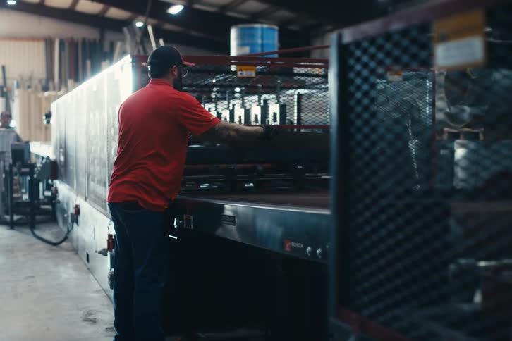 A factory worker in a red shirt feeds a large sheet of material into a manufacturing machine inside a warehouse