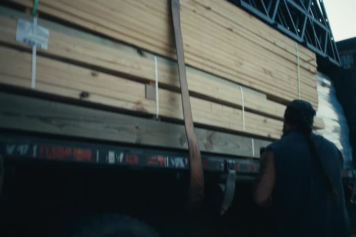 A blue-collar truck driver throws a tie-down strap over a load of lumber and trusses on his flatbed truck in slow motion