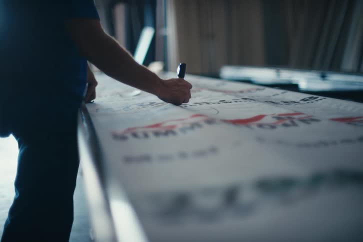 A male worker in a blue shirt carefully cuts a large printed sign on a workbench in a brightly lit workshop