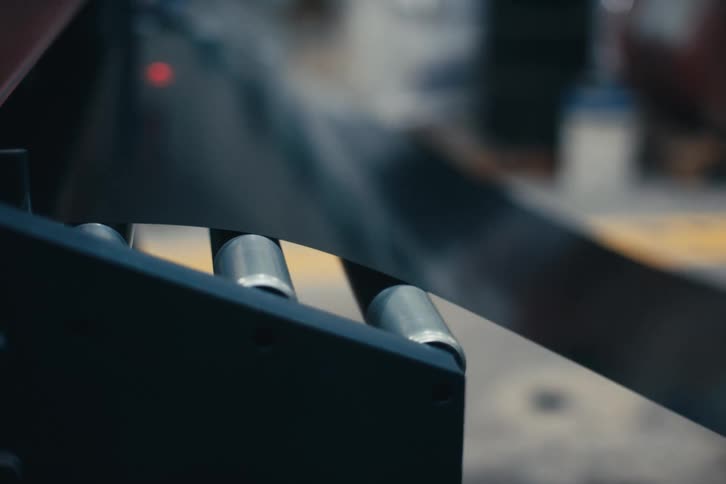 A sheet of thin, dark metal glides smoothly over rollers on a conveyor belt in an industrial factory setting