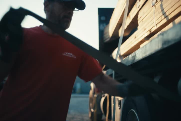 A truck driver in a red shirt tightens down a ratchet strap to secure a load of lumber onto a flatbed truck in cinematic slow motion