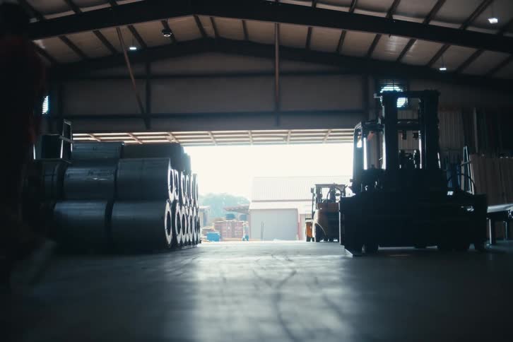 Two silhouetted workers walk through a large, dimly lit industrial warehouse past stacks of metal coils and a forklift