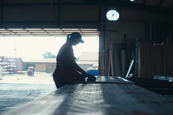 Silhouette of a female laborer sanding a long beam in an industrial warehouse with an open bay door