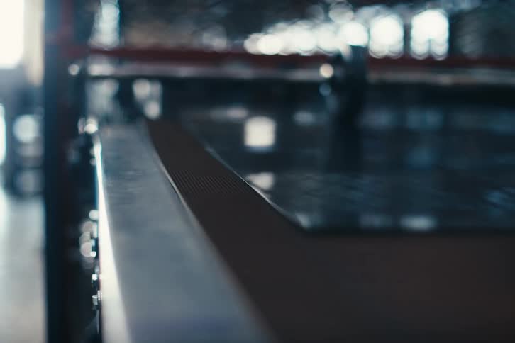 An automated manufacturing machine processes a sheet of glossy black material on a conveyor belt, with the camera pulling focus along its edge