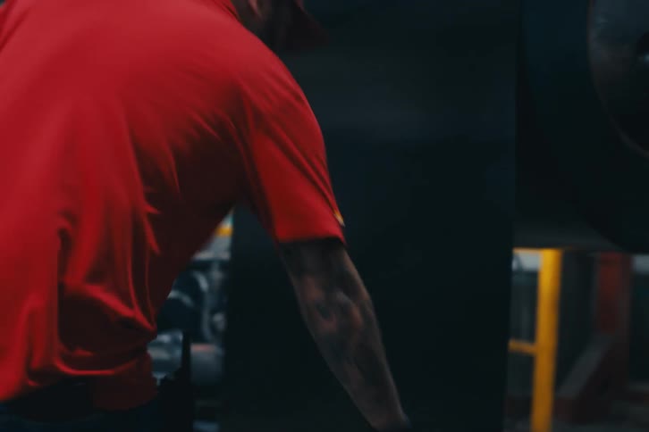 A factory worker with tattoos on his arm handles a large piece of sheet metal in slow motion inside an industrial warehouse