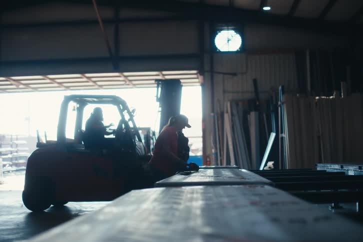 Silhouetted workers operate machinery and sand metal beams in a brightly lit industrial warehouse