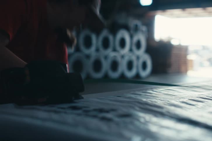 A factory worker in a dimly lit warehouse meticulously uses a banding tool to strap a large wrapped product for shipping