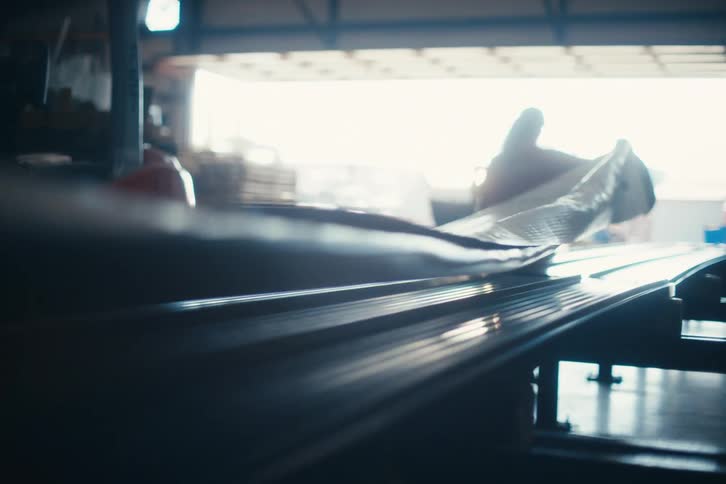 A long manufactured product moves along a conveyor belt in a cinematic, brightly lit warehouse