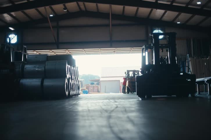 Low angle shot from inside a dark industrial warehouse looking out at forklifts and workers in the bright yard