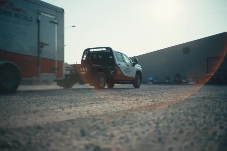 A work truck with a flatbed tows a large trailer across a gravel lot in slow motion during a sunny day