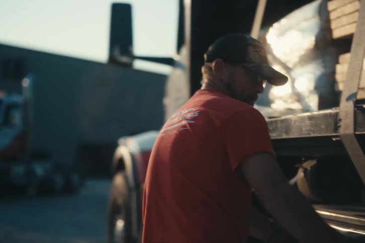 A truck driver in a red shirt and hat uses a winch to tighten straps and secure a load on a flatbed semi-truck at golden hour