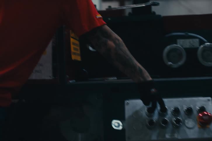 A male factory worker in a red polo and hat walks through a warehouse to operate the control panel of a large piece of machinery