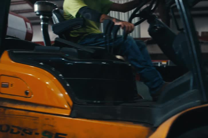 A forklift driver transports a large metal coil through an industrial warehouse as other workers walk by