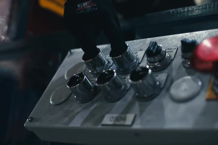 A factory worker's hand in a safety glove presses two silver switches on a machine's control console