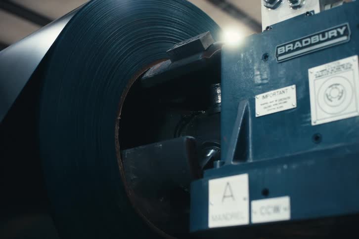 A close-up shot of a large roll of sheet metal loaded onto a Bradbury industrial machine in a factory setting