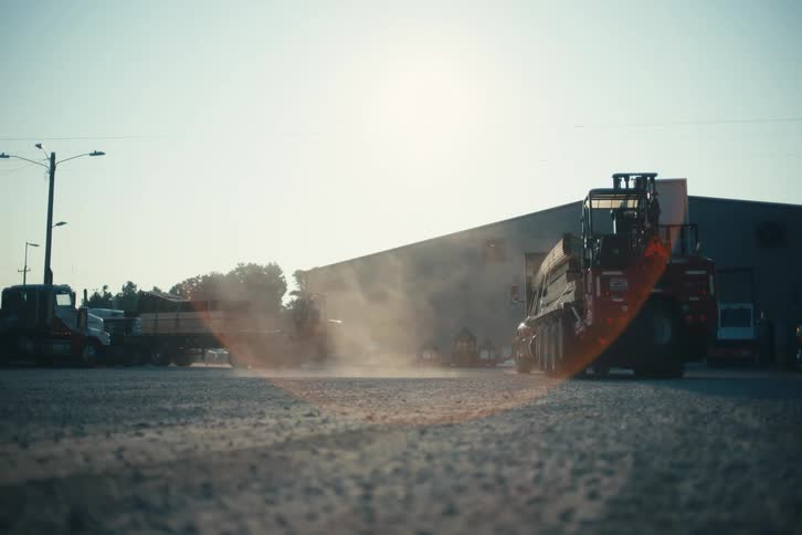 A large truck loaded with materials pulls away from an industrial yard, kicking up dust in the bright sunlight