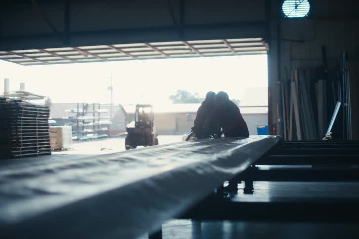 A team of silhouetted workers handle a long, wrapped product on a production line inside a sunlit factory