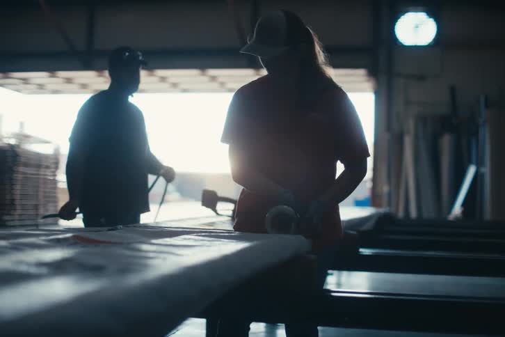 Two silhouetted factory workers tape and prepare a large package for shipping in a dimly lit warehouse