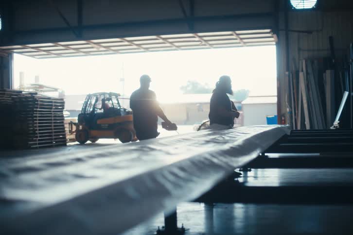 Workers in a manufacturing warehouse are silhouetted by the bright sun as they wrap a long product for shipping