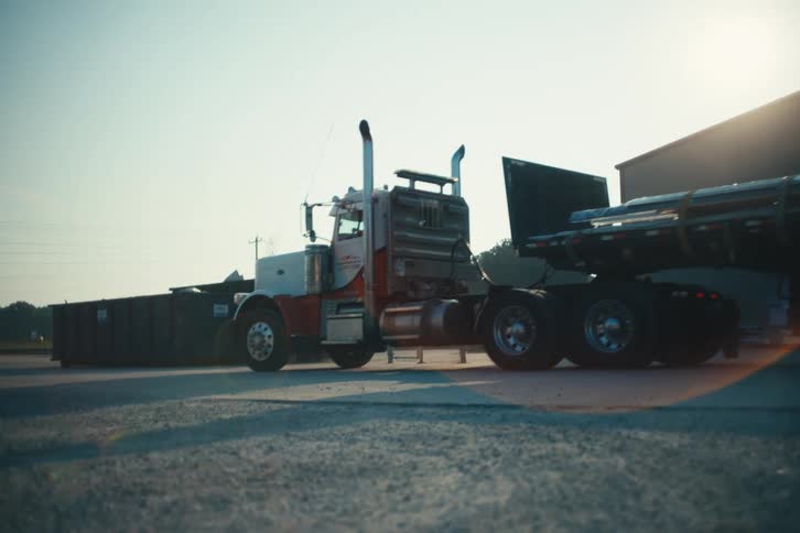 A large semi-truck slowly pulls a loaded flatbed trailer out of an industrial warehouse yard at sunrise