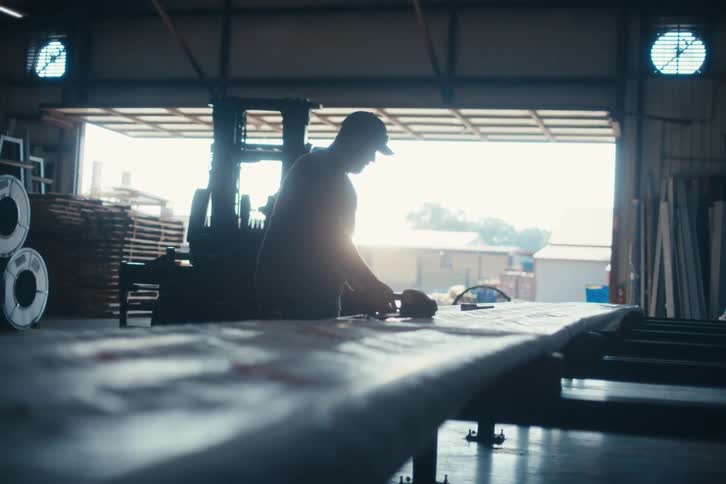 A silhouetted worker uses a tool on a long piece of material in an industrial warehouse with a bright open door
