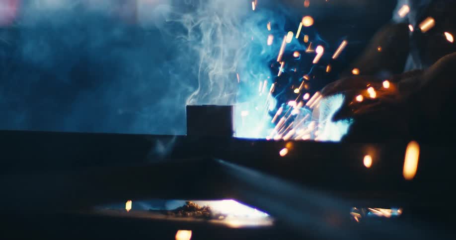 Cinematic slow-motion close-up of welding, where bright sparks fly and blue smoke billows in a dark industrial setting