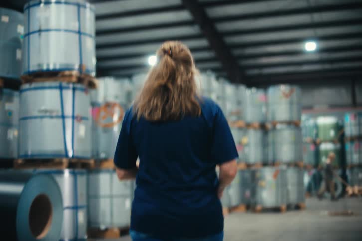 A female warehouse worker walks through a large factory floor filled with stacked metal coils