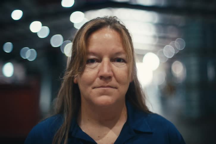 A serious female factory worker in a blue uniform slowly looks up at the camera in a brightly lit warehouse