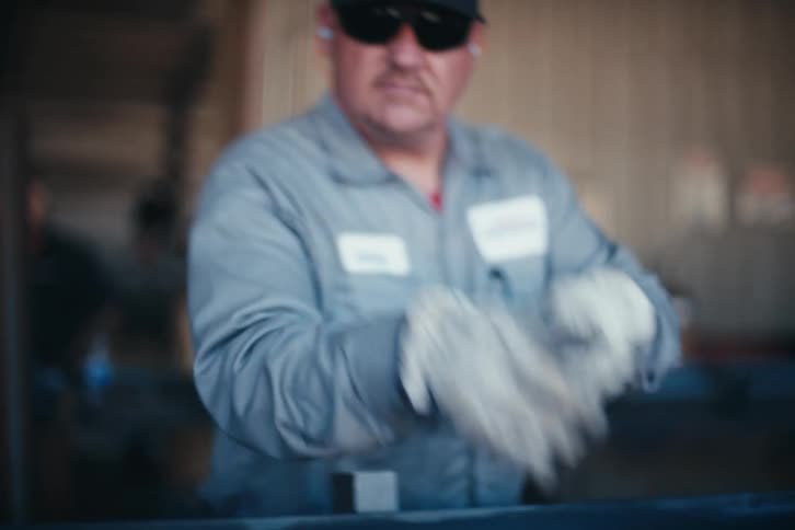 A factory worker in a grey uniform and gloves cleans a metal frame and then playfully aims two metal parts at the camera