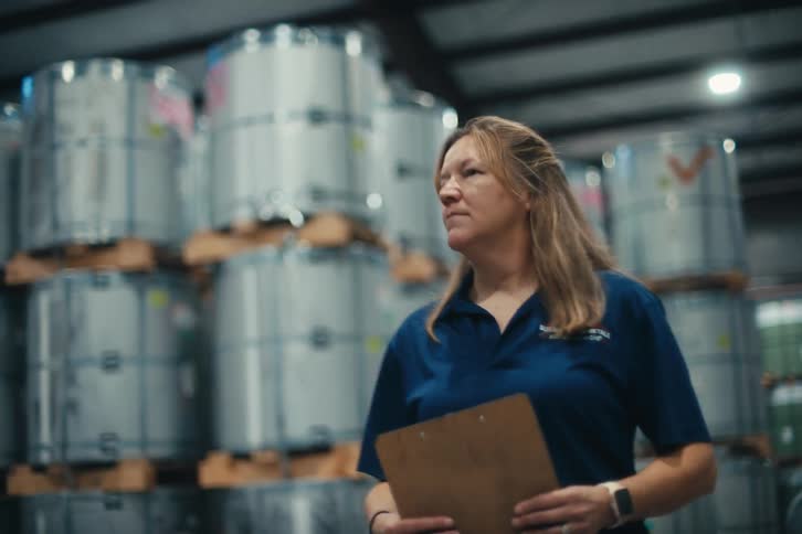 A determined female supervisor walks through an industrial warehouse with a clipboard, inspecting large metal coils in slow motion