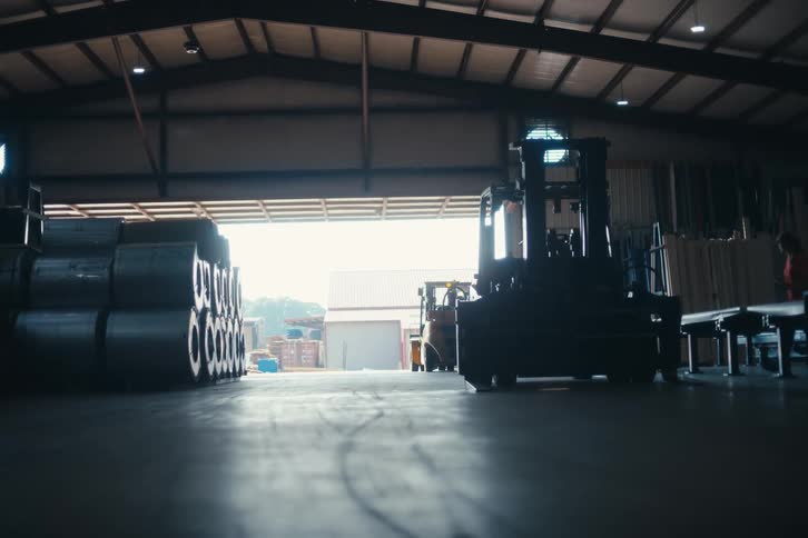 A low-angle view from inside a silhouetted warehouse shows a forklift and stacked metal rolls against the bright light of an open bay door as a worker walks by