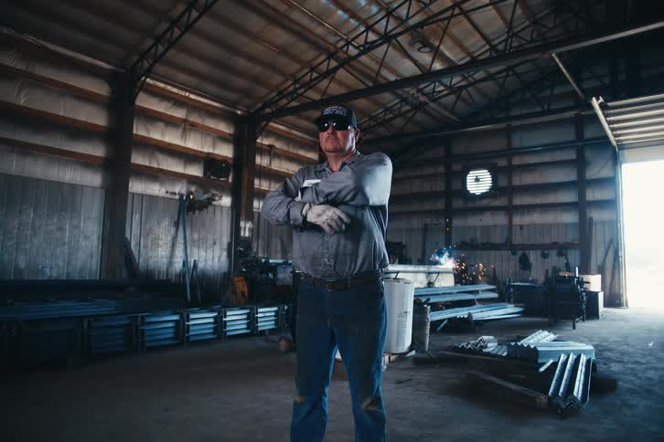 A confident metalworker stands proudly in his workshop while welding sparks fly in the background