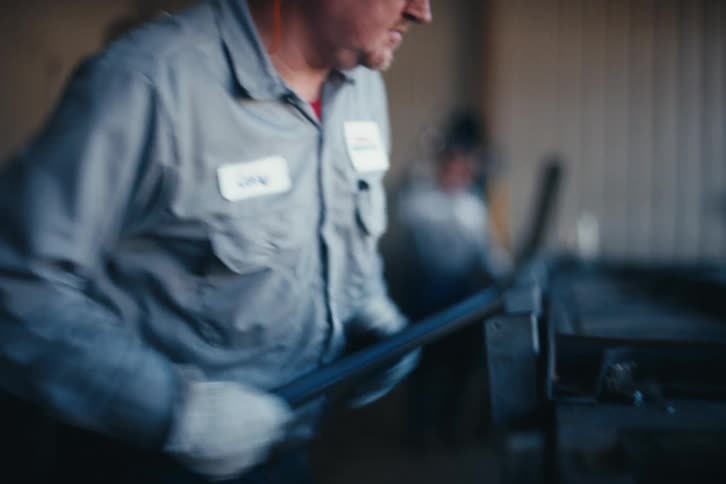 A male factory worker wearing gloves and safety gear smooths the edges of a metal piece in a workshop