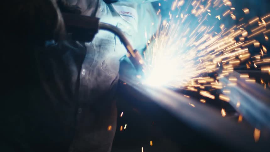 A welder in a protective mask works on a metal beam, creating a shower of bright sparks in slow motion
