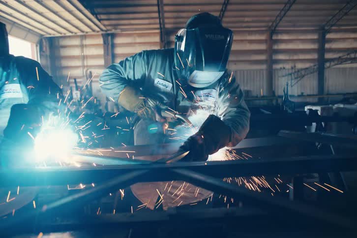 Two welders in protective gear work together on a metal frame, creating a shower of sparks in a cinematic slow-motion reveal