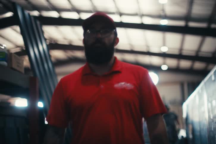 A bearded factory worker in a red shirt and hat walks through an industrial warehouse in slow motion